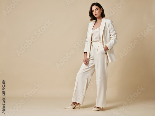 A woman posing in a cream colored oversized blazer and matching wide leg trousers with a hand in her pocket against a neutral studio backdrop