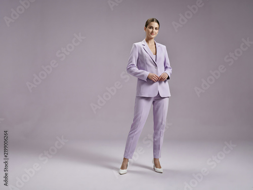 A woman with dark hair wearing a light purple blazer and matching trousers with white heels standing against a soft light purple background
