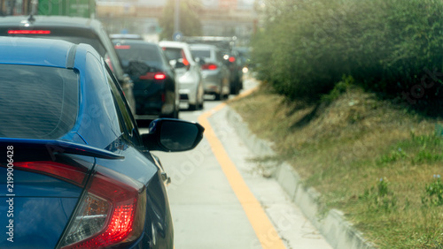 Blue car navigates through a slow line of traffic on an urban road during daytime. Side road with nature of green grass and small trees. Blazing sunlight on the streets of Thailand.