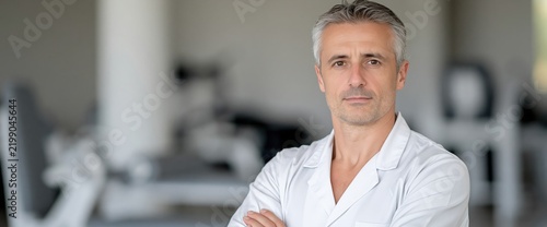 Physiotherapist with salt and pepper hair stands with crossed arms inside a modern rehabilitation center, looking forward with a confident and reassuring expression.