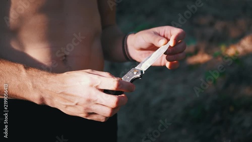 Shirtless man examines the sharp blade of a folding pocket knife outdoors. Warm afternoon sunlight highlights his hands and muscular torso against a soft forest background.