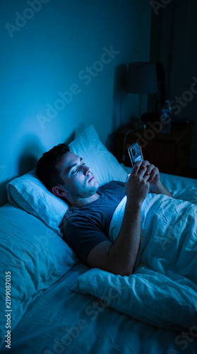 Young man lying in bed at night using smartphone in a dark bedroom