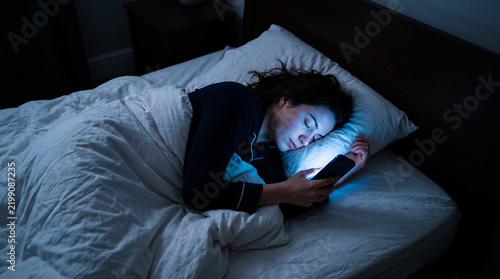 Young woman falling asleep in bed at night while holding a glowing smartphone