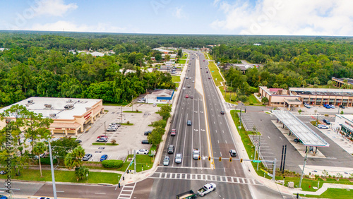 Aerial view of a busy multi-lane highway intersection with surrounding suburban commercial businesses and dense green tree line background busy road traffic flow suburban landsc
