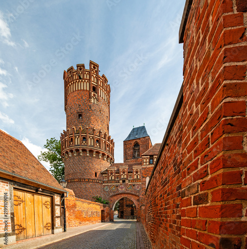 Historic medieval brick gate with round tower, coats of arms and surrounding brick walls along a cobblestone street, photographed in landscape format.