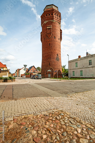 Historic brick water tower rising above a residential street with mixed paving and old cobblestones, photographed in portrait format.