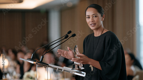 Maori business leader giving acceptance speech holding crystal trophy, indigenous cultural background acknowledged with subtle pendant, corporate boardroom awards dinner, New