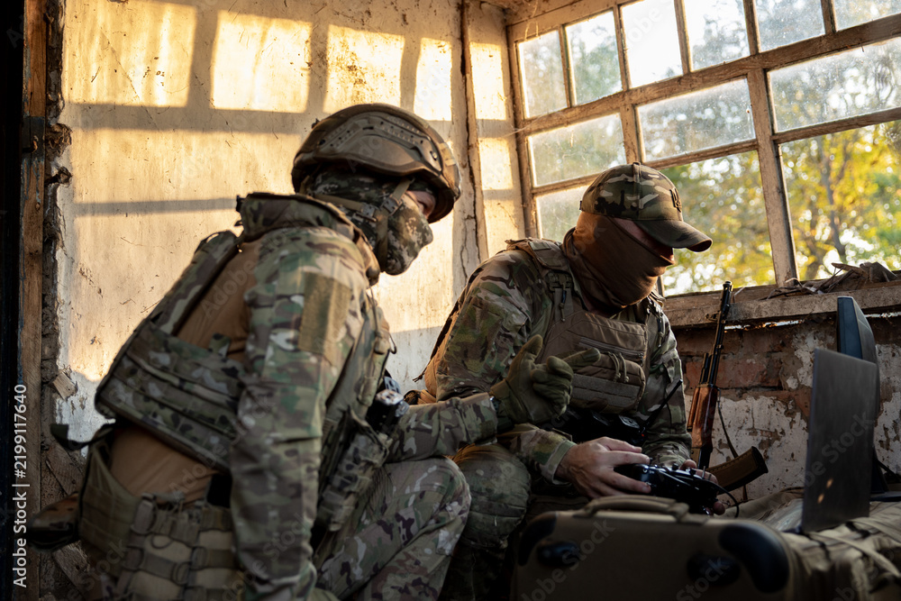 Obraz premium Soldiers operate a drone control console. Military personnel are carrying out a combat mission at a position in an abandoned house.