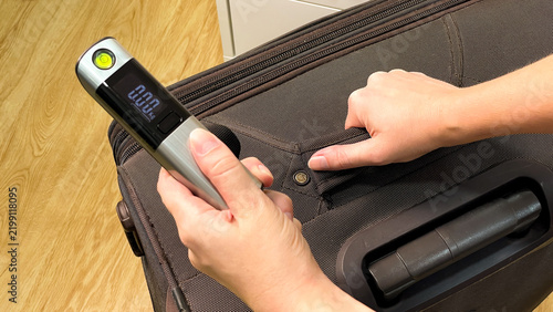 A woman's hands check a large travel suitcase using a compact electronic scale. Checking the luggage weight helps estimate how heavy it is in advance.