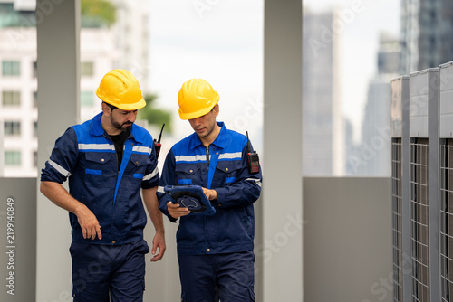 Industrial engineer inspecting equipment with coworker on rooftop, wearing protective helmet and uniform during building maintenance work in urban setting