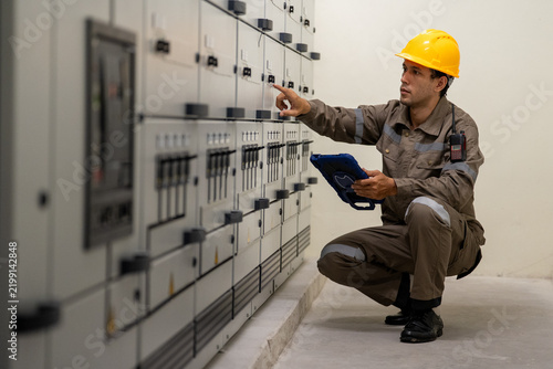 Industrial engineer inspecting electrical control panel in protective workwear, using tablet inside clean utility room with focused attention and caution