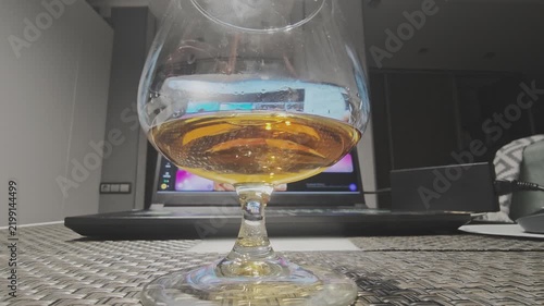 Golden amber cognac swirling in a snifter glass. The laptop screen reflects on the elegant glassware, creating a sophisticated ambiance