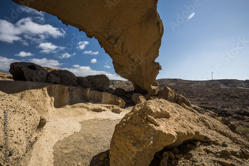Natural rock formation resembling a massive stone arch or bridge in an arid and rocky landscape under a clear blue sky with some clouds. Tenerife, Spain.