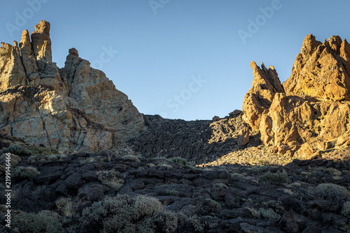 Dramatic rock formations rise above a volcanic landscape with dark lava fields and sparse vegetation. The scene is illuminated by natural light in Tenerife, Canary Islands, Spain.