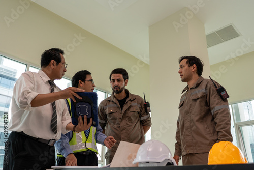 Engineer industrial team discussing safety equipment in bright workshop, with protective helmet and high visibility vest visible during focused professional briefing