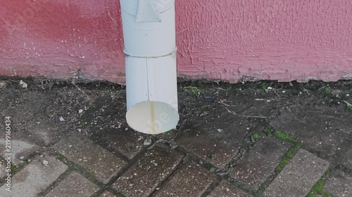 Rainwater cascading down a white downspout onto damp brick paving below. A detailed view of the drainage system positioned at the bottom of a crimson painted building facade
