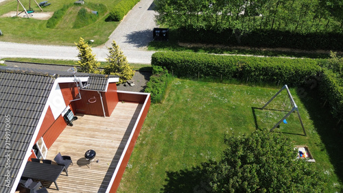 Aerial view of a traditional red wooden holiday home in Bork Havn, West Jutland. The property features a wooden deck with patio furniture, a green lawn with a swing set and sandbox, and a gravel path