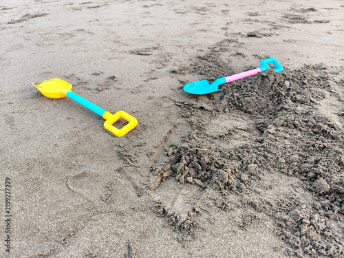 Two colorful plastic toy shovels lay on the beach sand. One shovel was yellow with a blue handle, while the other was blue with a pink handle.