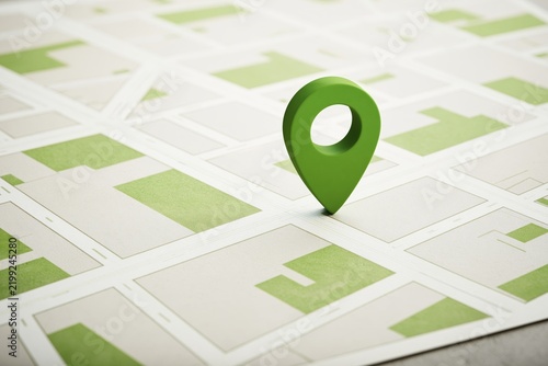 Green three-dimensional location marker standing upright on a printed street map showing abstract urban grid layout with beige and olive colored blocks and intersecting roads under bright even