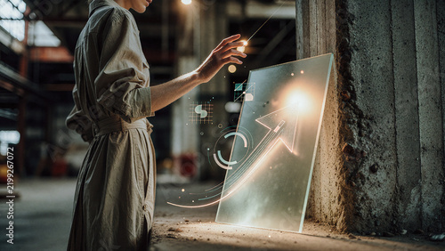 Woman in tan linen attire interacts with glowing digital graphics on a transparent glass panel within an industrial warehouse. Warm rim lighting highlights her profile and the upward arrow icon.