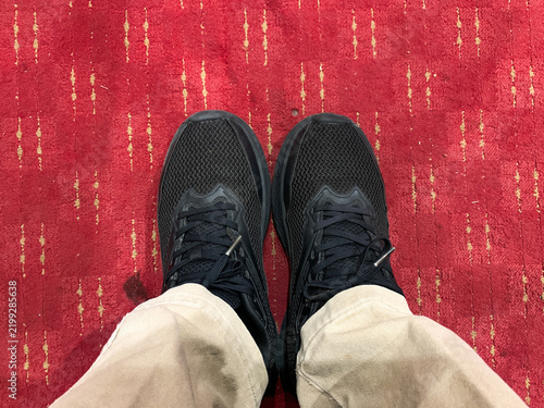 Top-down photo of black sneakers and beige pants standing on a red patterned carpet, evoking casual travel, waiting, everyday movement and relaxed indoor footwear in textured interior flooring.
