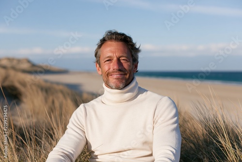 Smiling man seated on sandy dunes in a white turtleneck for coastal lifestyle portrait and calm beach background design