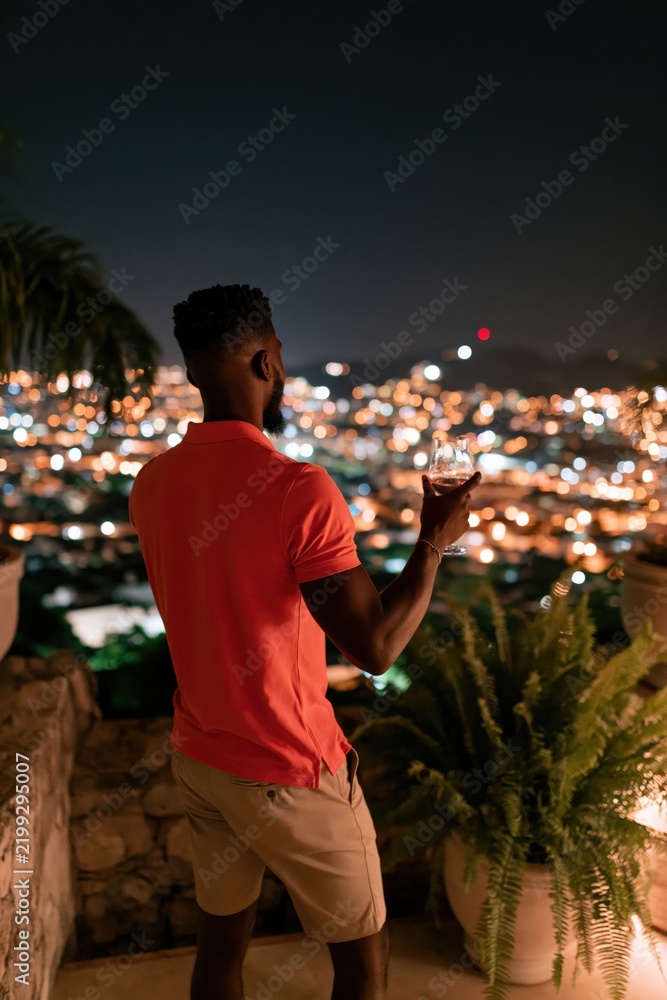 Fototapeta premium Man on terrace holding a wine glass overlooking city lights at night for lifestyle background and evening relaxation banner