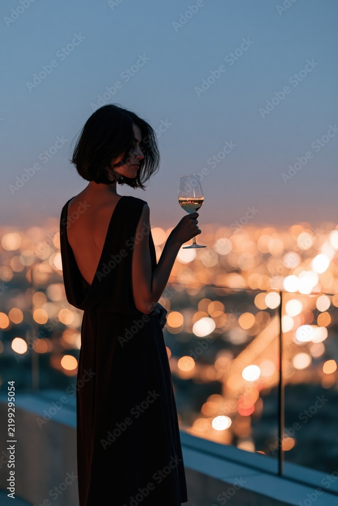 Obraz premium Elegant woman on rooftop balcony holding a wine glass against bokeh city lights for evening background and banner design
