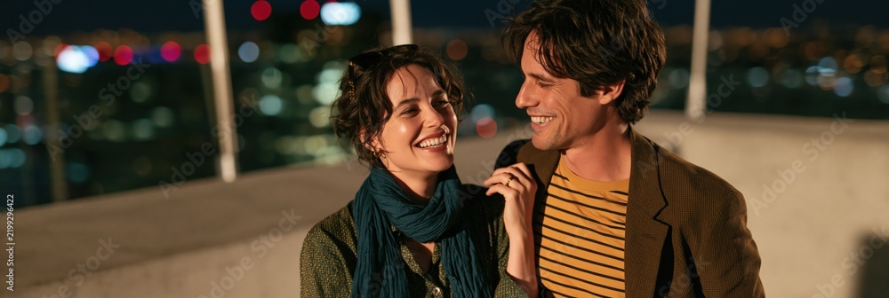 Fototapeta premium Smiling couple on a rooftop at night city background for romantic banner and evening lifestyle wallpaper design