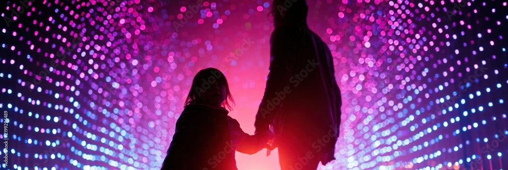 Fototapeta premium Silhouette of parent and child holding hands walking through a neon led light tunnel with pink and purple bokeh for synthwave background poster banner