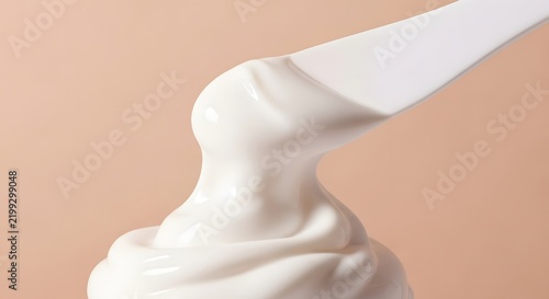 A white creamy substance being scooped by a spatula against a soft peach background, highlighting its smooth texture.