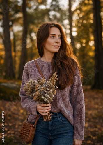 A woman in a cozy knit sweater holds a bouquet of dried wildflowers on a gentle walk through an autumn forest. Surrounded by golden leaves, soft sunlight, and tall trees with seasonal vibe