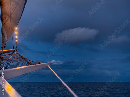 Fotografie The bowsprit and its net stretch out over dark water at night, lit by small bulbs, distant harbour lights on the horizon