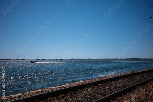 Boats move across the water near a railway track on a clear day with blue sky