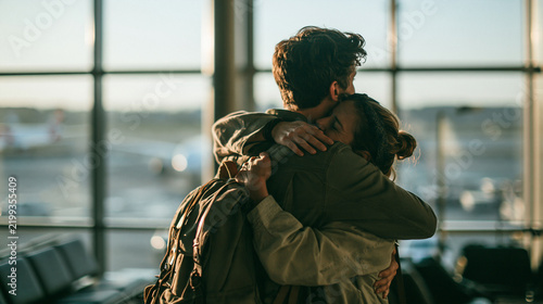 A Fond Farewell at the Airport: Two travelers share an emotional hug at the airport, silhouetted against the large windows overlooking the tarmac. The warm sunlight casts long shadows.