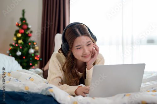 Smiling young Asian woman relaxing on bed with headphones using laptop while watching video at home during Christmas holidays.