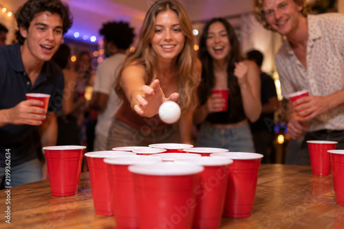 Low table-level view of red cups arranged for beer pong as a young woman takes her shot and friends react around the game table.