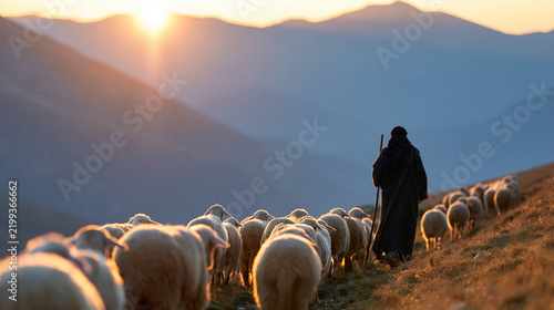 Faceless shepherd guiding a flock of sheep across a vast mountain landscape at sunrise, parable of pastoral care, faith and spiritual leadership, with copy space