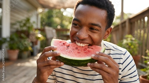Happy African American man eating fresh slice of watermelon on wooden patio outdoors