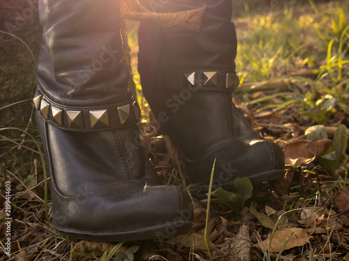 Close-up of black leather witch-style boots with metallic studs among fallen autumn leaves, illuminated by warm golden backlight in a woodland Halloween setting.