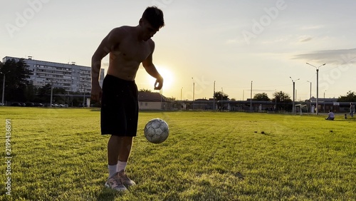 Young man juggling soccer ball on stadium at sunset. Professional footballer kicking ball at green field. Sportsman practicing tricks at meadow with sunlight at background. Freestyle football