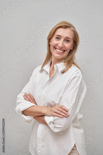 Blonde Woman Standing With Arms Crossed And Smiling In White Shirt Against Plain Gray Studio Background.