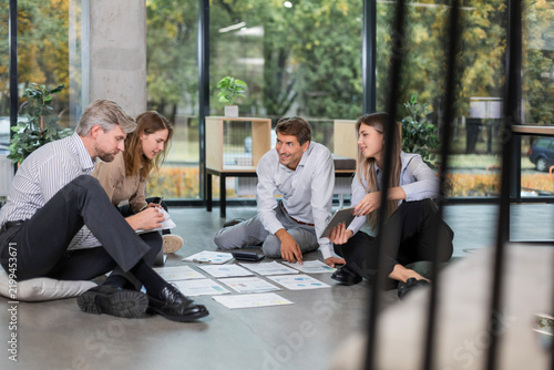 Group of business people making researchers during brainstorming session sitting on floor talking , team of creative co-workers using technology and papers planning startup collaborating on strategy