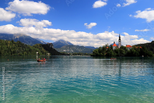 Lake Bled, Slovenia: Wide scenic view of the crystal clear turquoise lake water with tourist rowboats, the famous island with church, and mountains under a blue sky.
