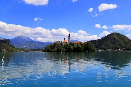 Lake Bled, Slovenia: Wide scenic view of the crystal clear turquoise lake water with tourist rowboats, the famous island with church, and mountains under a blue sky.