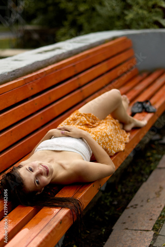 Joyful beautiful girl resting on a bright orange park bench outdoors, wearing a white strapless top and floral skirt, looking at the camera with a gentle smile on a damp day.