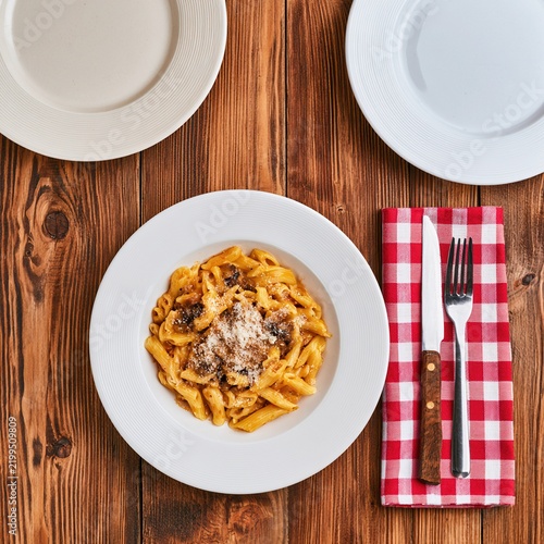 A plate of penne pasta with sauce and grated cheese, placed on a rustic wooden table with empty plates and cutlery on a red and white checkered napkin.
