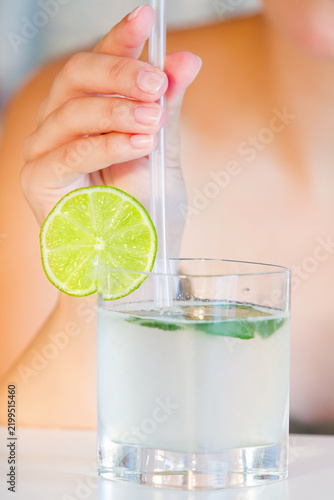 Woman is holding a clear glass filled with a light-colored drink, garnished with a lime slice and a straw.