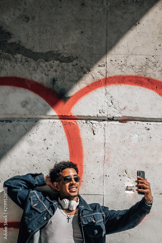 Young african american man smiling confidently while taking a selfie with his smartphone against an urban graffiti wall, expressing joy and lifestyle energy in an outdoor street