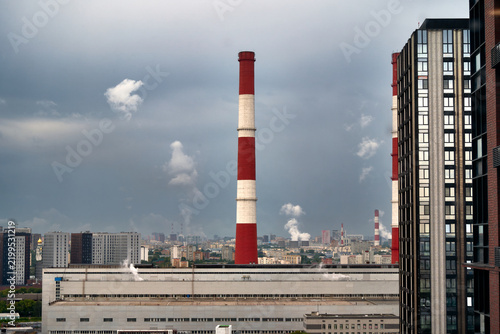 Power plant smokestacks stand beside modern city buildings. Steam rises beneath a cloudy sky over the skyline. Industrial energy and pollution shape urban development.